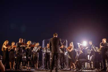 Orchestra Performing Live Concert Under Blue Night Sky – VYB STUDIOS Full orchestra performing an outdoor live concert under a deep blue night sky with stage lighting.